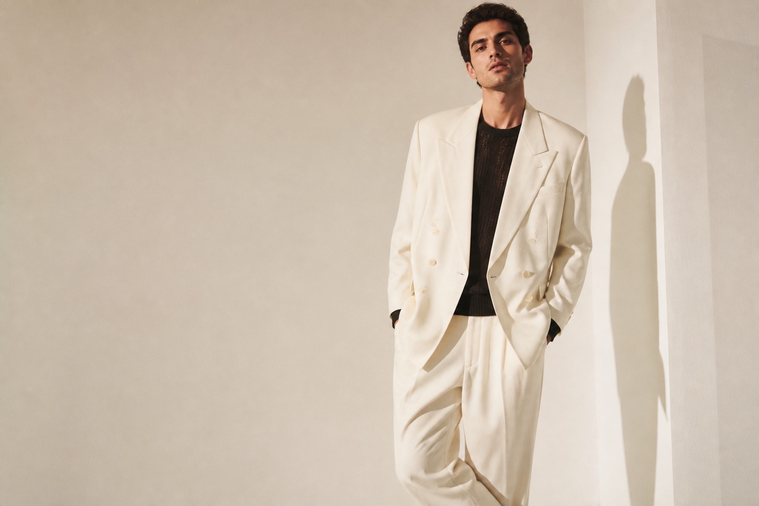 Man in an oversized cream suit and black knit standing in a warm ivory studio